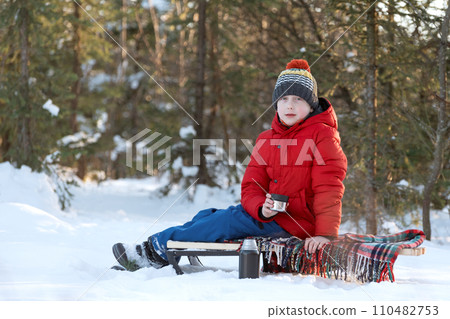 Boy in warm puffy jacket and knitted hat sits on sleigh with cup of hot cocoa tea or hot chocolate in winter in forest. Boy in warm puffy jacket and knitted hat sits on sleigh with cup of hot cocoa tea or hot chocolate in winter in forest. 110482753