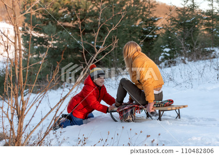 Boy pushes his mothers or older sisters sled in snow-covered forest. Have fun outside for family holiday in winter. 110482806