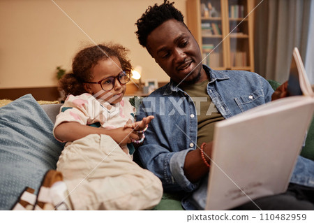 Medium long shot of little African American girl wearing glasses at home with dad reading stories 110482959