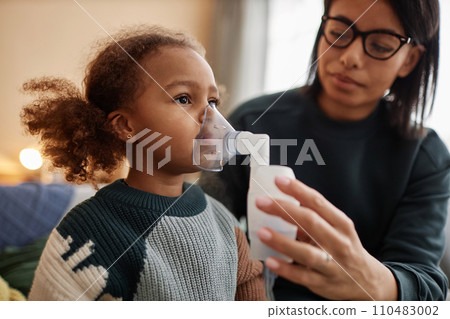 Medium close up shot of little African American girl inhaling medicine through nebulizer while defocused mom holding face mask on daughters face Medium close up shot of little African American girl inhaling medicine through nebulizer while defocused mom holding face mask on daughters face 110483002