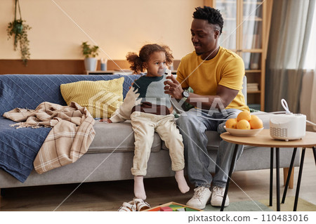 Full shot of smiling African American dad helping ill little daughter with inhalation through nebulizer while sitting on big sofa at home 110483016