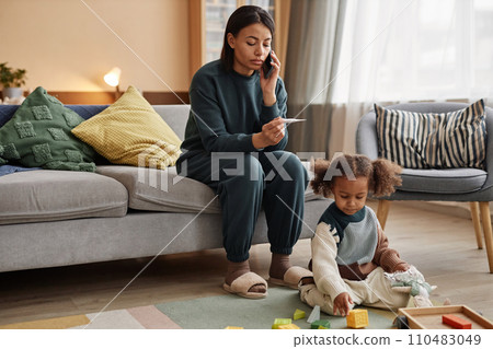 Full length shot of African American mother calling pediatrician with thermometer in hand while daughter playing with toys on floor at home 110483049