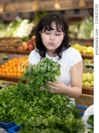 Girl of asian appearance carefully chooses fresh parsley in supermarket Girl of asian appearance carefully chooses fresh parsley in supermarket 110483250