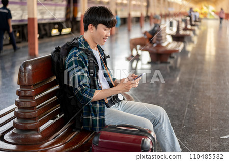 A man tourist is using his phone while sitting at a bench on a platform at a railway station. 110485582