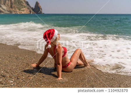 Female beach Santa hat wave coast. beach relaxation seaside. A woman in a red swimsuit enjoying her time on the beach, sits on the sand and being covered by a wave. 110492870