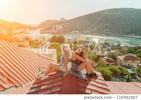 Two women sitting on a red roof, enjoying the view of the town and the sea. Rooftop vantage point. In the background, there are several boats visible on the water, adding to the picturesque scene. 110492907