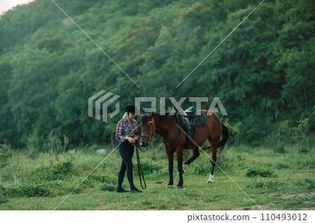 Happy blonde with horse in forest. Woman and a horse walking through the field during the day. Dressed in a plaid shirt and black leggings. 110493012