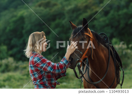 Happy blonde with horse in forest. Woman and a horse walking through the field during the day. Dressed in a plaid shirt and black leggings. 110493013