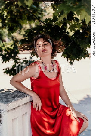 woman in a red silk dress and a bandage on her head smiles against the background of the leaves of a tree. She is leaning on the coop and looking into the camera. Vertical photo. woman in a red silk dress and a bandage on her head smiles against the background of the leaves of a tree. She is leaning on the coop and looking into the camera. Vertical photo. 110493014