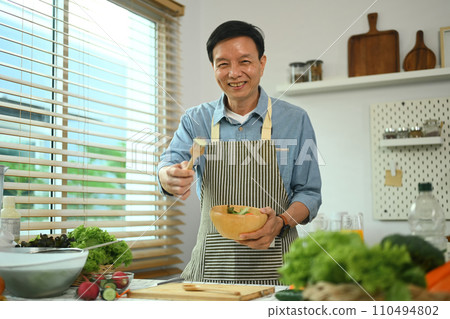 Positive middle age man in apron holding a bowl of salad and smiling at camera. Healthy eating concept Positive middle age man in apron holding a bowl of salad and smiling at camera. Healthy eating concept 110494802