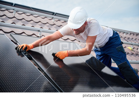 Worker building solar panel system on rooftop of house. Man engineer in helmet installing photovoltaic solar module outdoors. Alternative, green and renewable energy generation concept. 110495536