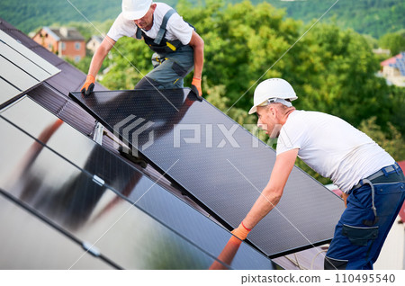 Workers building solar panel system on rooftop of house. Two men installers in helmets installing photovoltaic solar module outdoors. Alternative, green and renewable energy generation concept. Workers building solar panel system on rooftop of house. Two men installers in helmets installing photovoltaic solar module outdoors. Alternative, green and renewable energy generation concept. 110495540
