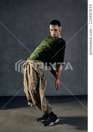 Athletic male with tattooed body, earrings, beard. Dressed in khaki t-shirt, jumpsuit, black sneakers. Dancing on gray background. Dancehall, hip-hop 110497614