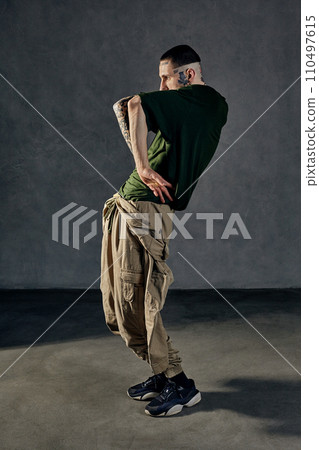 Athletic man with tattooed body, earrings, beard. Dressed in khaki t-shirt, overalls, black sneakers. Dancing on gray background. Dancehall, hip-hop 110497615