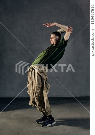Flexible fellow with tattooed body, earrings, beard. Dressed in khaki t-shirt, jumpsuit, black sneakers. Dancing, gray background. Dancehall, hip-hop 110497616