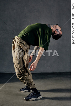 Graceful guy with tattooed body, earrings, beard. Dressed in khaki t-shirt, overalls, black sneakers. Dancing on gray background. Dancehall, hip-hop 110497628