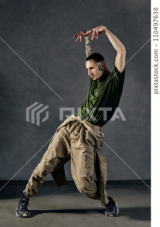 Fellow with tattooed body, earrings, beard. Dressed in khaki t-shirt and jumpsuit, black sneakers. Dancing on gray background. Dancehall, hip-hop Fellow with tattooed body, earrings, beard. Dressed in khaki t-shirt and jumpsuit, black sneakers. Dancing on gray background. Dancehall, hip-hop 110497638