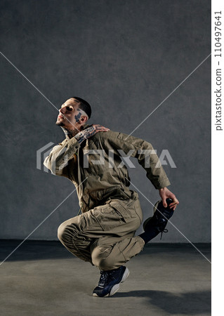 Extraordinary guy with tattooed body, earrings, beard. Dressed in khaki overalls and black sneakers. Dancing on gray background. Dancehall, hip-hop 110497641