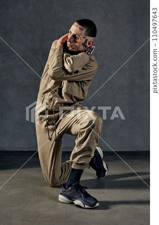 Unusual performer with tattooed body, earrings, beard. Dressed in khaki overalls and black sneakers. Dancing on gray background. Dancehall, hip-hop 110497643