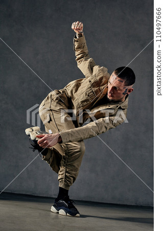 Attractive male with tattooed body, earrings, beard. Dressed in khaki jumpsuit and black sneakers. Dancing on gray background. Dancehall, hip-hop Attractive male with tattooed body, earrings, beard. Dressed in khaki jumpsuit and black sneakers. Dancing on gray background. Dancehall, hip-hop 110497666