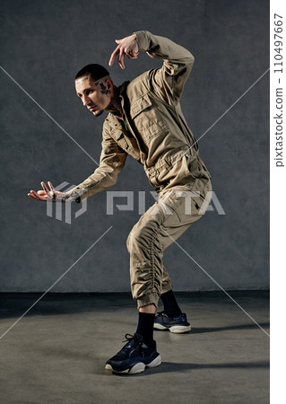 Flexible performer with tattooed body, earrings, beard. Dressed in khaki overalls and black sneakers. Dancing on gray background. Dancehall, hip-hop Flexible performer with tattooed body, earrings, beard. Dressed in khaki overalls and black sneakers. Dancing on gray background. Dancehall, hip-hop 110497667