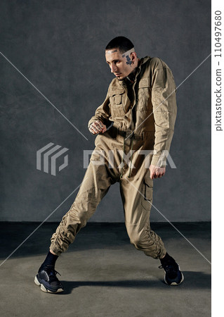 Active performer with tattooed body, earrings, beard. Dressed in khaki overalls and black sneakers. Dancing on gray background. Dancehall, hip-hop 110497680