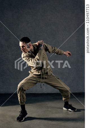 Stately man with tattooed body and face, earrings, beard. Dressed in khaki overalls and black sneakers. Dancing on gray background. Dancehall, hip-hop Stately man with tattooed body and face, earrings, beard. Dressed in khaki overalls and black sneakers. Dancing on gray background. Dancehall, hip-hop 110497713