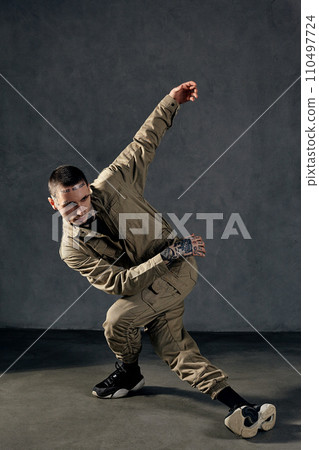 Good-looking fellow with tattooed body, earrings, beard. Dressed in khaki jumpsuit, black sneakers. Dancing on gray background. Dancehall, hip-hop Good-looking fellow with tattooed body, earrings, beard. Dressed in khaki jumpsuit, black sneakers. Dancing on gray background. Dancehall, hip-hop 110497724
