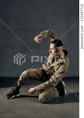 Athletic guy with tattooed body and face, earrings, beard. Dressed in khaki overalls, black sneakers. Dancing on gray background. Dancehall, hip-hop 110497728