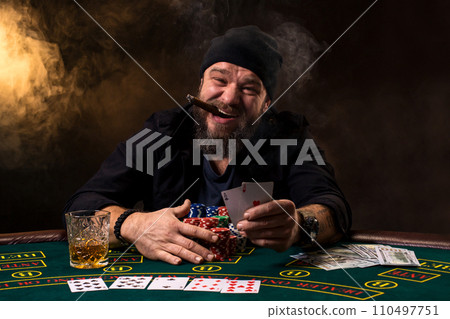 Bearded man with cigar and glass sitting at poker table in a casino. Gambling, playing cards and roulette. 110497751