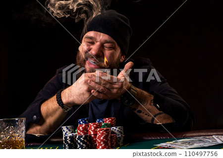 Bearded man with cigar and glass sitting at poker table in a casino. Gambling, playing cards and roulette. 110497756