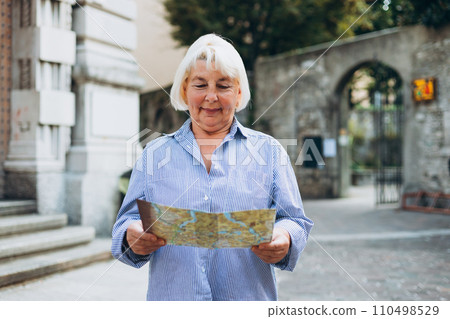 Mature blonde woman tourist looking at city map at street. Cheerful 60s woman traveling abroad in summer. Travel and active lifestyle concept Mature blonde woman tourist looking at city map at street. Cheerful 60s woman traveling abroad in summer. Travel and active lifestyle concept 110498529