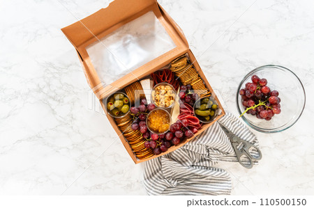 Flat lay. In the process of assembling, a woman expertly curates this charcuterie box, showcasing sliced meat, cheese, crackers, and grapes, all neatly arranged and packaged in a brown gifting box. 110500150