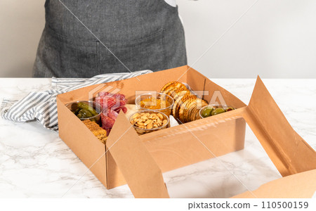 In the process of assembling, a woman expertly curates this charcuterie box, showcasing sliced meat, cheese, crackers, and grapes, all neatly arranged and packaged in a brown gifting box. 110500159