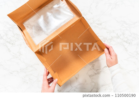 Flat lay. In the process of assembling, a woman expertly curates this charcuterie box, showcasing sliced meat, cheese, crackers, and grapes, all neatly arranged and packaged in a brown gifting box. 110500190