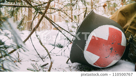 Corpsman Helmet Lies Near Fallen Soldier. Metal Helmet Of United States Army Infantry Medic Soldier At World War Ii. Re-enactor Dressed As USA Soldier Medic In Winter Season. Army Of World War II Corpsman Helmet Lies Near Fallen Soldier. Metal Helmet Of United States Army Infantry Medic Soldier At World War Ii. Re-enactor Dressed As USA Soldier Medic In Winter Season. Army Of World War II 110500767