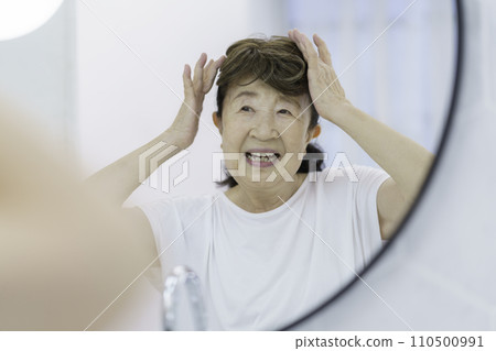 Smiling senior woman putting on a partial wig in front of the mirror 110500991