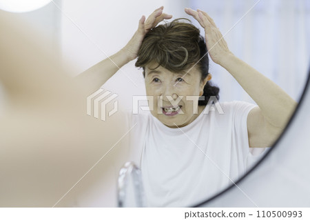 Smiling senior woman putting on a partial wig in front of the mirror 110500993