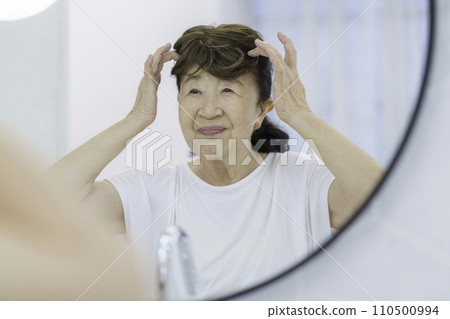 Smiling senior woman putting on a partial wig in front of the mirror 110500994