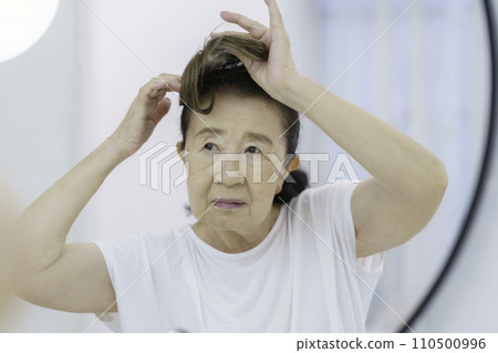 A senior woman with a troubled face putting on a partial wig in front of the mirror 110500996