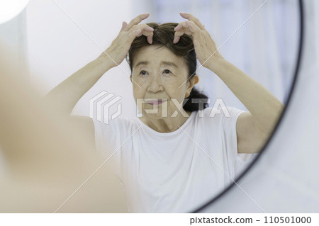 A senior woman with a troubled face putting on a partial wig in front of the mirror 110501000