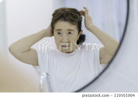 A senior woman with a troubled face putting on a partial wig in front of the mirror 110501004