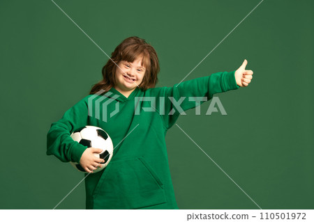 Happy smiling teen girl with down syndrome standing with football ball against green studio background. Sportive life 110501972