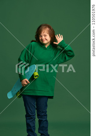 Teen girl with down syndrome wearing cozy hoodie, standing with skateboard against green studio background 110501976