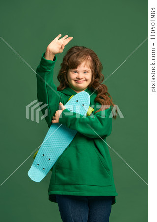 Teen girl with down syndrome wearing cozy hoodie, standing with skateboard against green studio background Teen girl with down syndrome wearing cozy hoodie, standing with skateboard against green studio background 110501983
