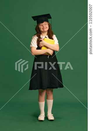 Inclusive education campaign. Girl with down syndrome standing with books, wearing graduation cap against green studio background 110502007