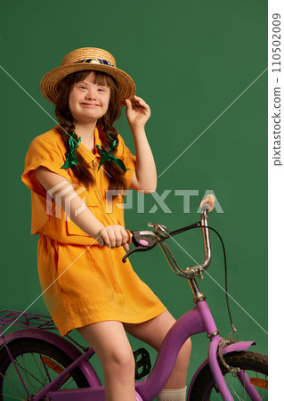 Happy teen girl with down syndrome wearing yellow suit, sitting on bicycle and having fun against green studio background Happy teen girl with down syndrome wearing yellow suit, sitting on bicycle and having fun against green studio background 110502009