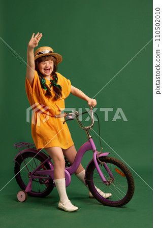 Happy teen girl with down syndrome wearing yellow suit, sitting on bicycle and having fun against green studio background 110502010