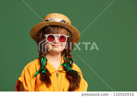Portrait of beautiful little girl with down syndrome wearing hat, sunglasses and yellow suit against green studio background 110502018