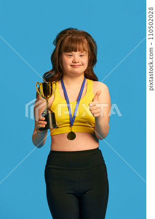 Promotion of equality and acceptance. Teen girl with down syndrome standing with medal and trophy against blue studio background 110502028